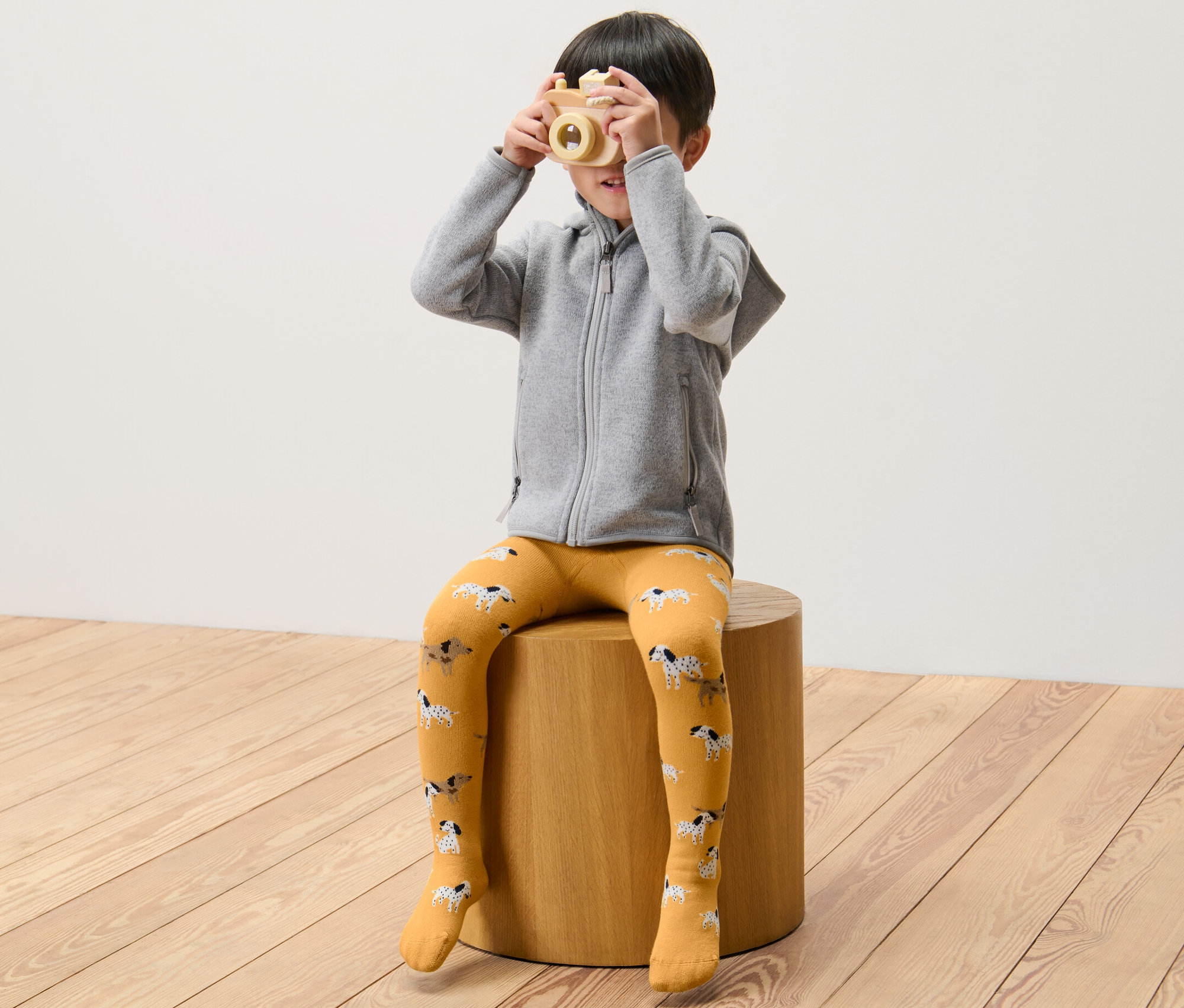 Un garçon est assis sur un tabouret et tient un appareil photo en bois devant son visage. Il porte 2 collants thermiques pour enfant, vert et jaune.