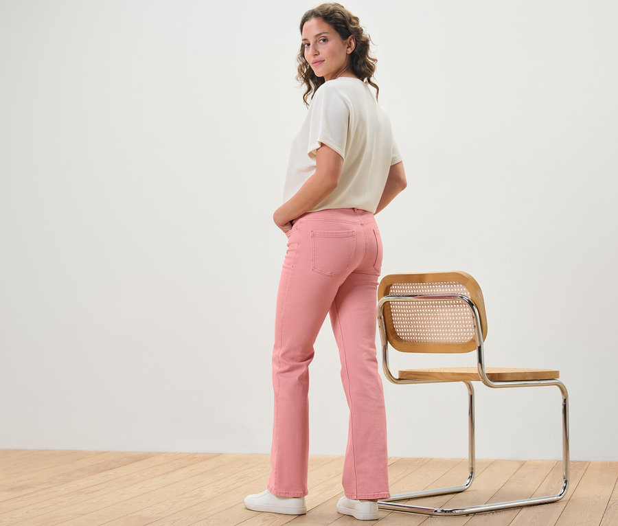 Femme posant en jean bootcut corail – Coupe « Lou » à côté d'une chaise.