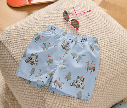 Un boardshort pour enfant avec un motif de chevalier posé sur un pouf, à côté d'un skateboard, de lunettes de soleil et d'un ballon.