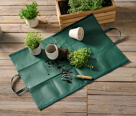 Un sac à déchets de jardin repose sur un plancher en bois à côté de plantes en pot et d'un arrosoir.