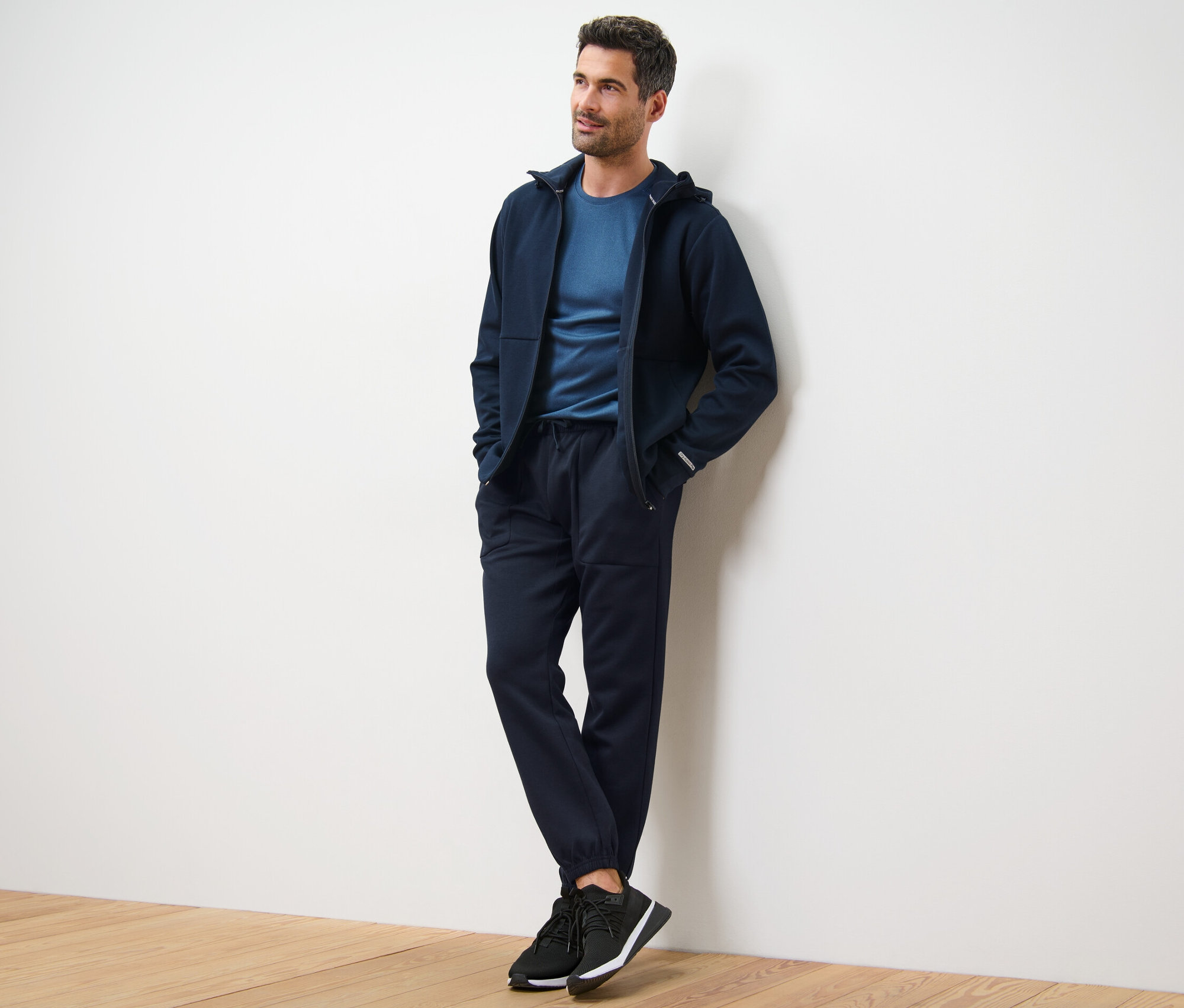 Un homme appuyé contre un mur porte un sweat à capuche bleu marine, un t-shirt et un pantalon technique en sweat.