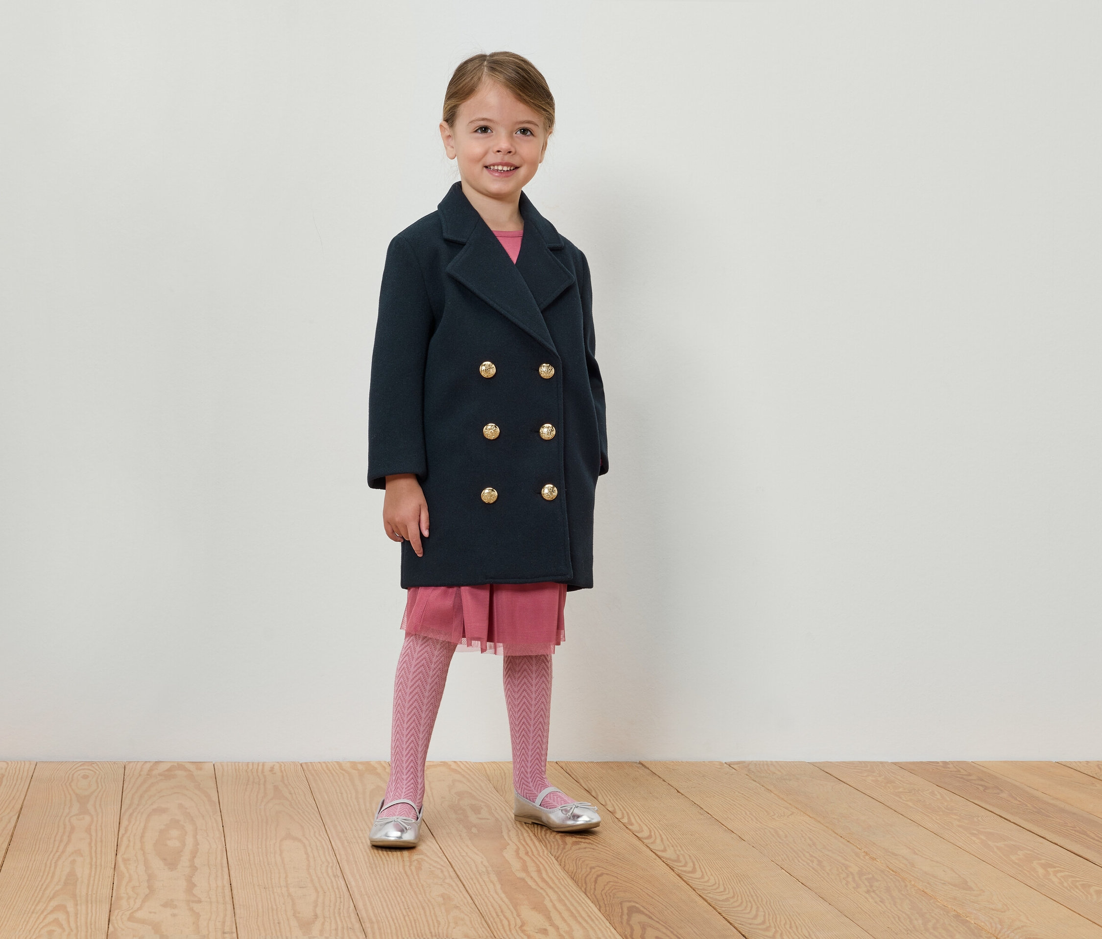 Fillette debout en manteau croisé bleu marine, robe rose et collants.