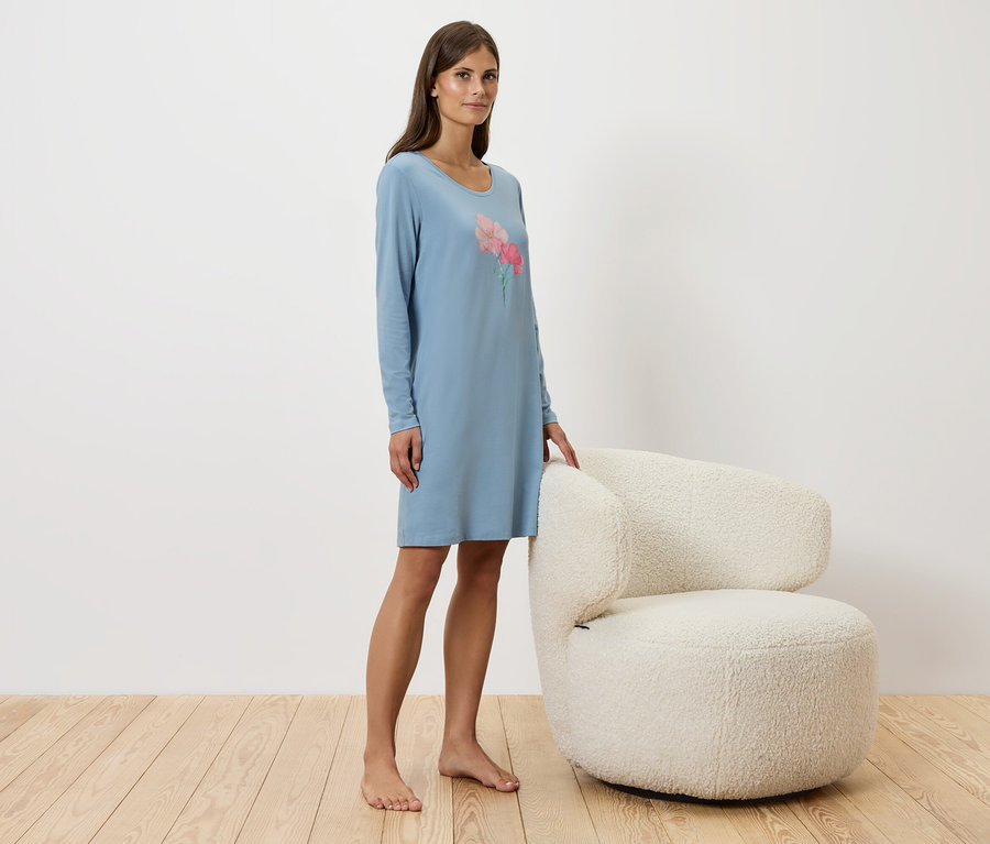 Une femme se tient à côté d'un fauteuil blanc et porte une chemise de nuit bleue.