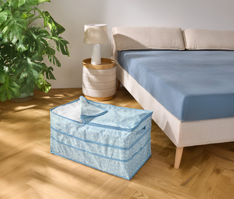 Sur un parquet, une boîte de rangement à motifs bleu clair est posée à côté d'un lit avec un drap bleu.