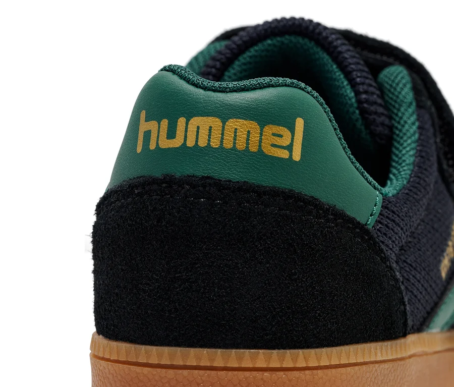 Gros plan d'une chaussure HUMMEL VM78 CPH JR avec du daim noir, du tricot bleu foncé et un logo vert.