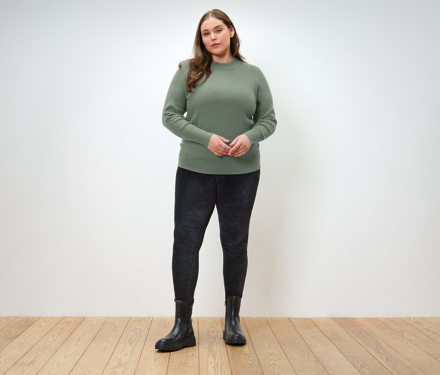 Une femme se tient sur un plancher en bois et porte un pull vert, un jean noir et des bottes noires.