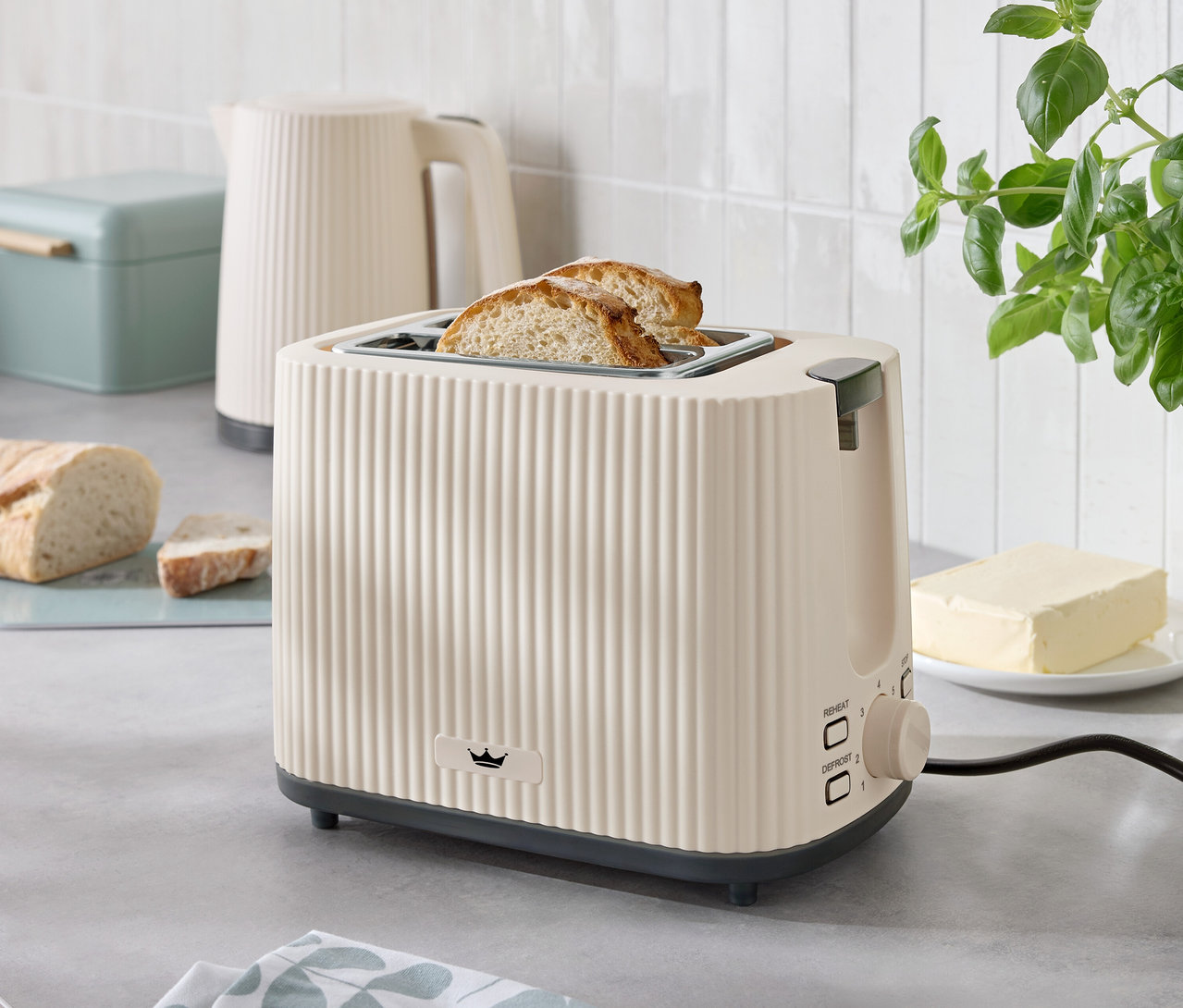 Deux tranches de pain grillé dépassent d'un grille-pain beige sur un comptoir de cuisine, à côté d'une bouilloire, d'un beurrier et d'une boîte à pain.