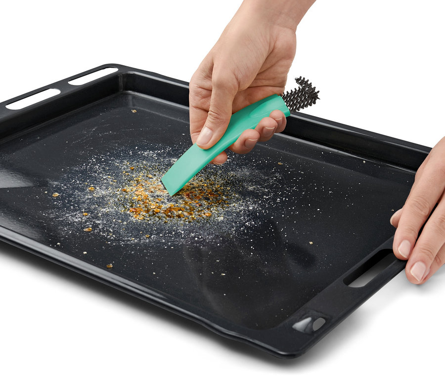 Une main nettoie une plaque de cuisson noire avec une brosse à four avec racloir.
