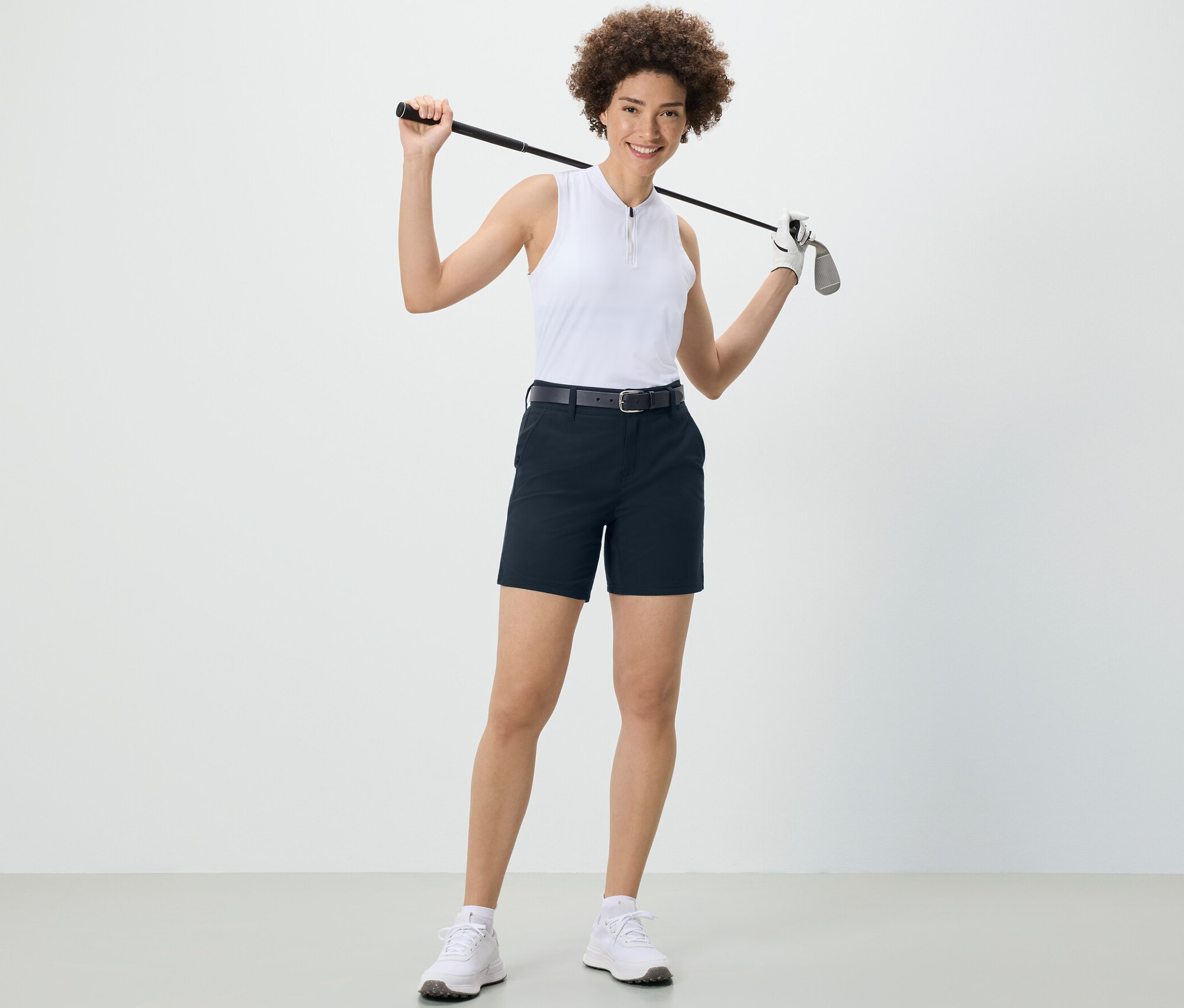 Une femme tient un club de golf sur ses épaules et porte un short technique en softshell bleu foncé.