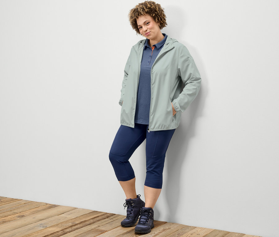 Une femme appuyée contre un mur porte un legging de trekking 3/4, un polo fonctionnel bleu foncé et une veste fonctionnelle pliable grise.