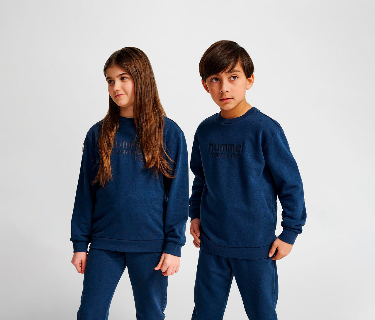Deux enfants se tiennent côte à côte et portent des sweat-shirts Hummel HMLJR Base bleus et des pantalons de survêtement bleus.