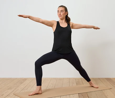 Femme en débardeur noir et leggings, debout sur un tapis de yoga, en position de guerrier II.