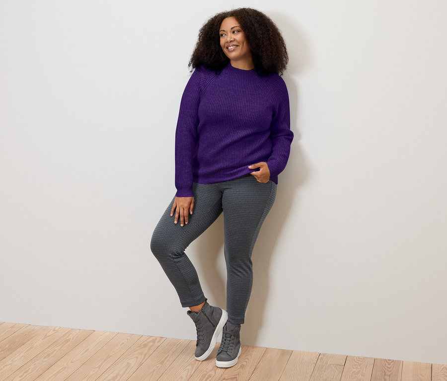 Femme posant en pull à col droit en grosse maille lilas, un jegging gris et des baskets.