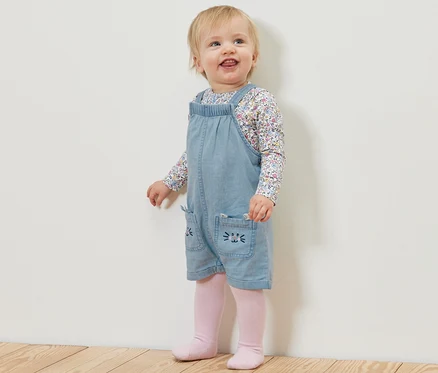 Un jeune enfant se tient sur un plancher en bois, portant une salopette en chambray pour enfant et un t-shirt à fleurs.
