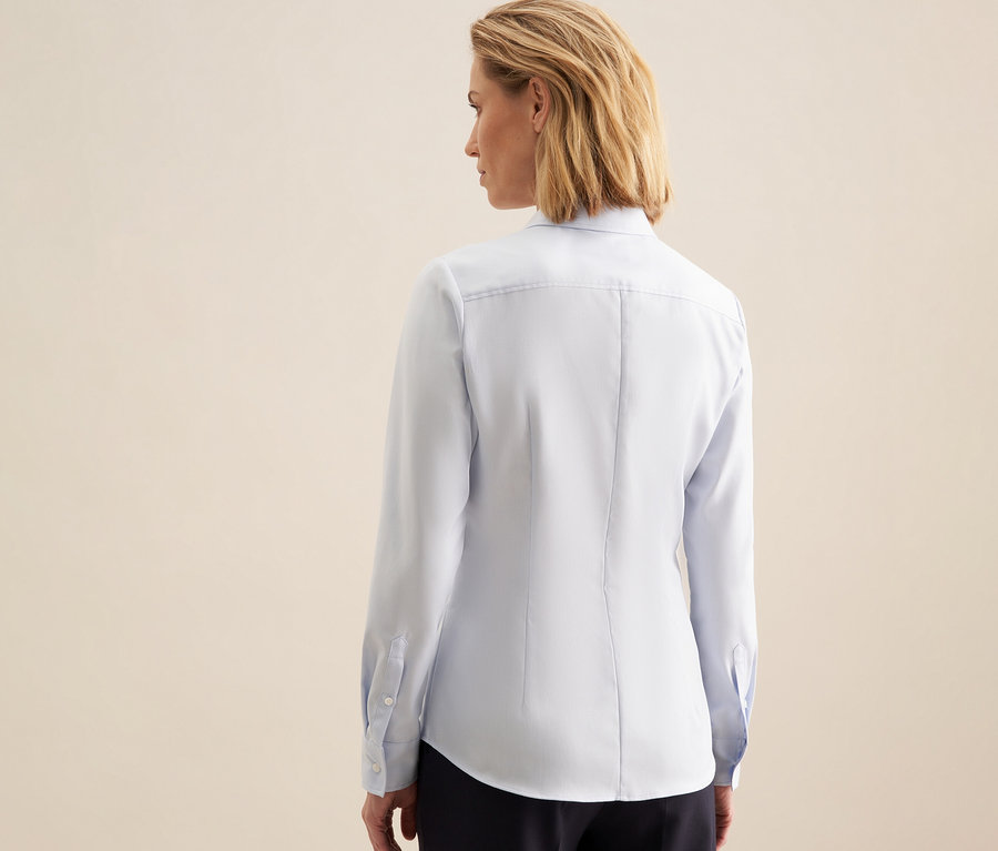 Femme de dos, la tête légèrement tournée vers la droite, portant un chemisier bleu clair Seidensticker et un pantalon foncé.
