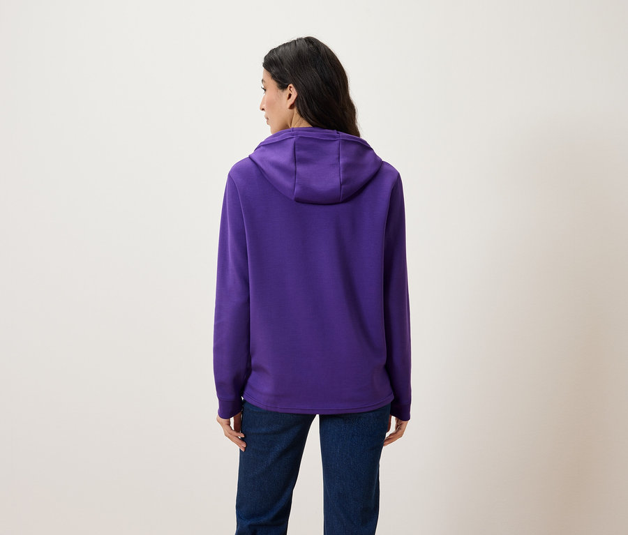 Une femme vue de dos porte un sweat à capuche violet et un jean.
