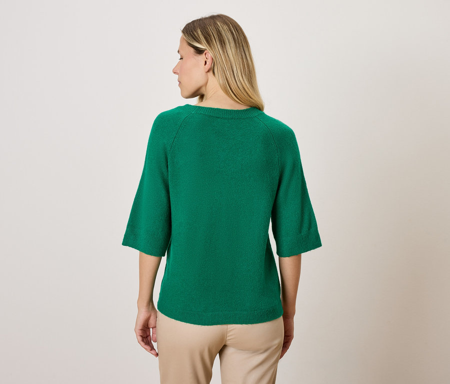 Une femme portant un pull vert à manches trois quarts et un pantalon beige, dos à la caméra.