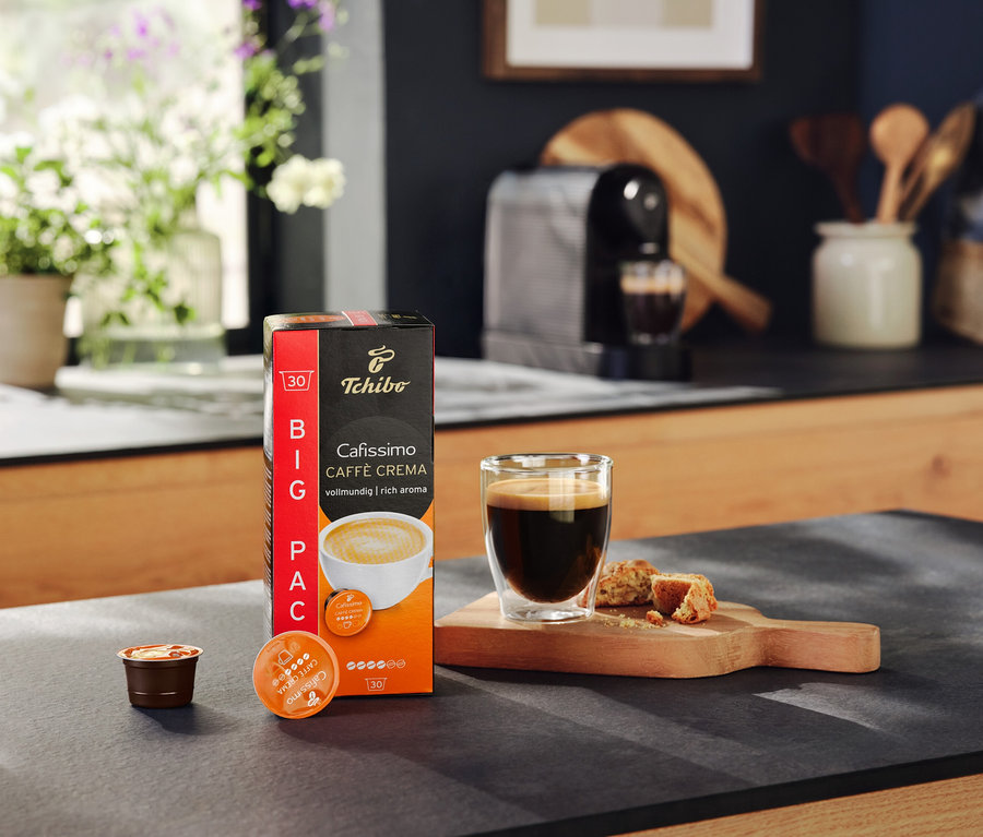 Un paquet de capsules Cafissimo Caffe Crema, un verre de café et des biscuits sur une table. À l'arrière-plan, une machine à café Cafissimo pure+ black.