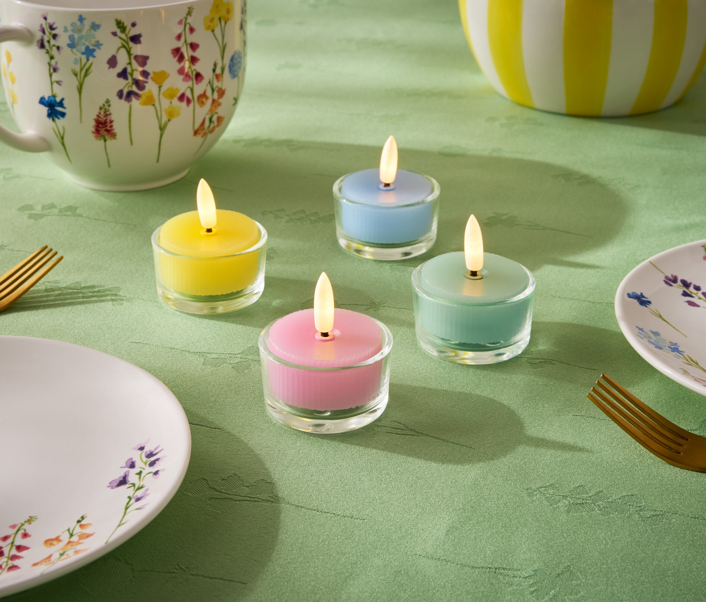 Sur une nappe verte, quatre petites bougies de couleurs pastel : jaune, bleu, vert et rose. On y trouve également une assiette, une tasse et un bol à motifs floraux, ainsi que deux fourchettes dorées.