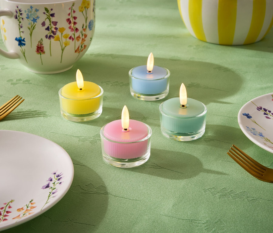 Sur une nappe verte, quatre petites bougies de couleurs pastel : jaune, bleu, vert et rose. On y trouve également une assiette, une tasse et un bol à motifs floraux, ainsi que deux fourchettes dorées.