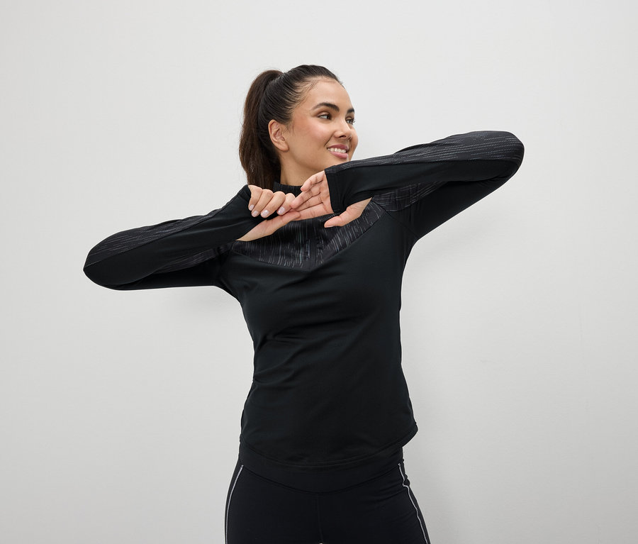 Une femme souriante en tenue de sport noire s'étire devant un fond blanc.