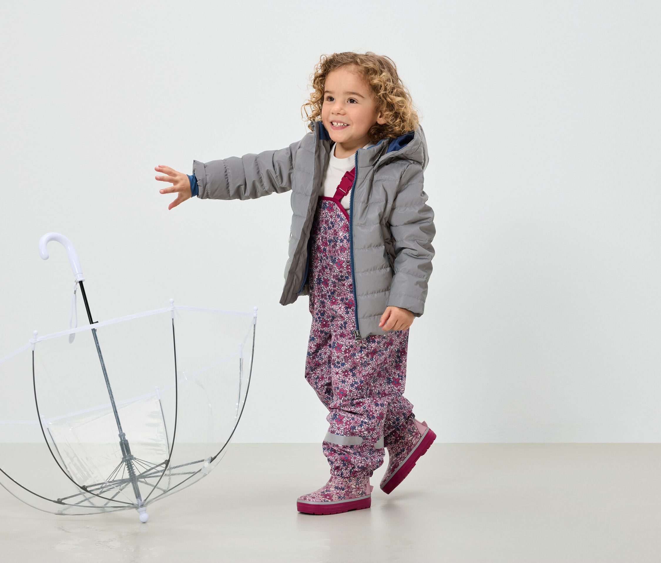 Une fillette souriante aux cheveux bouclés, vêtue d'une veste grise et d'un pantalon à fleurs, se tient à côté d'un parapluie transparent.