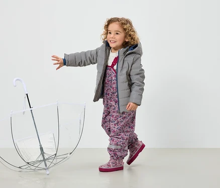 Une fillette souriante aux cheveux bouclés, vêtue d'une veste grise et d'un pantalon à fleurs, se tient à côté d'un parapluie transparent.