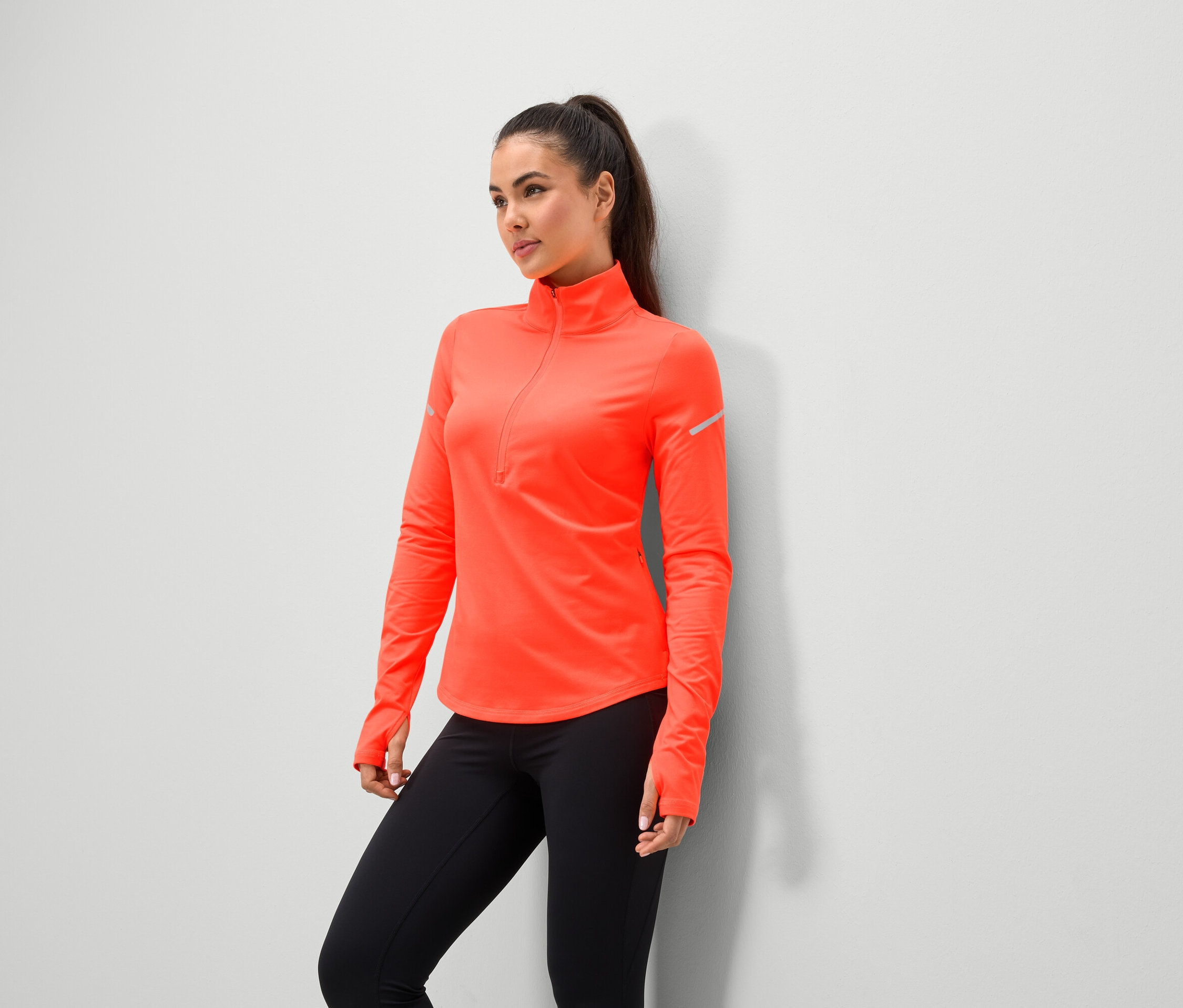 Une femme portant une veste de sport corail et des leggings noirs se tient contre un mur.