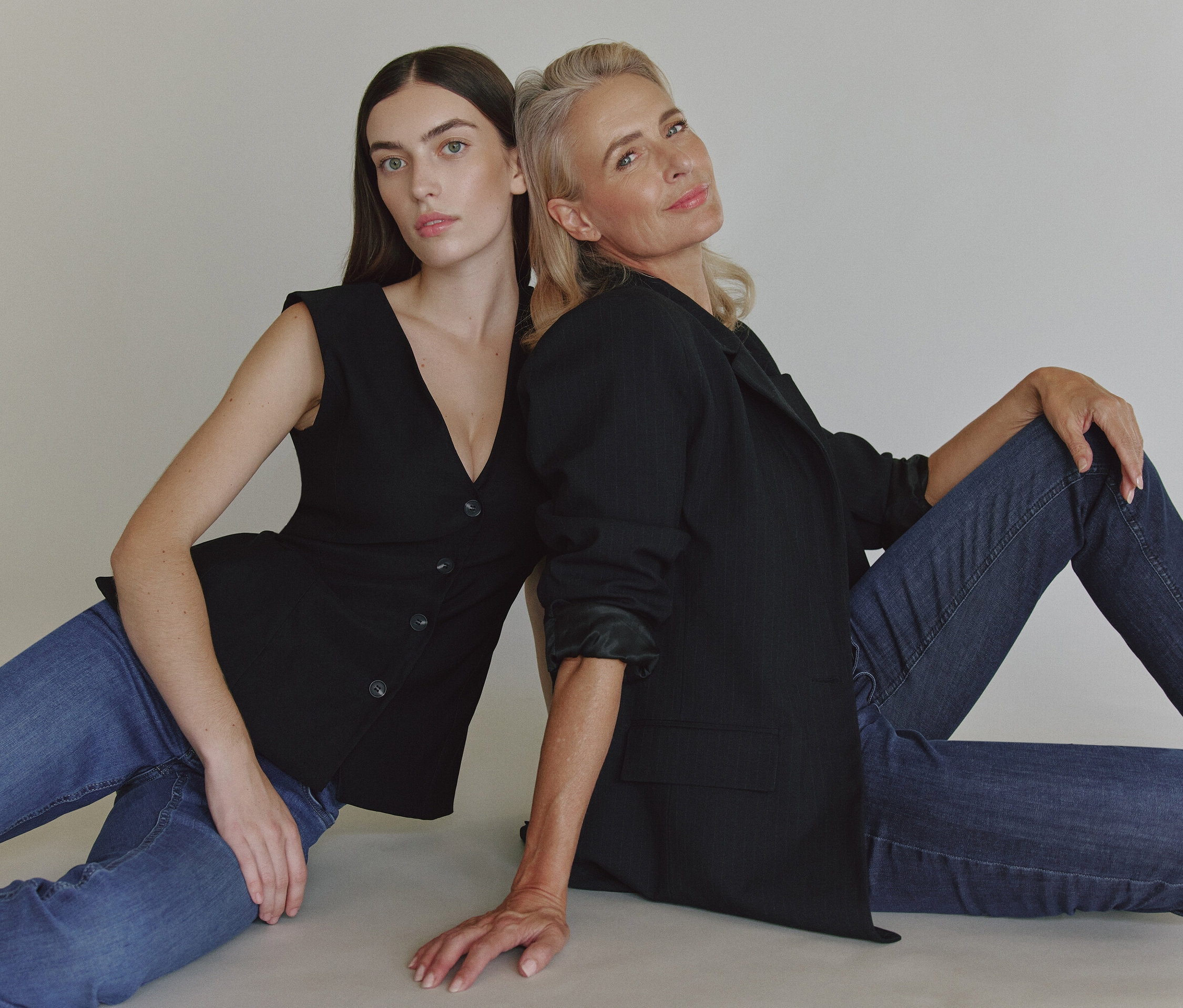 Deux femmes sont assises dos à dos sur le sol. L'une porte une veste noire et un jean, l'autre un gilet noir et un jean.