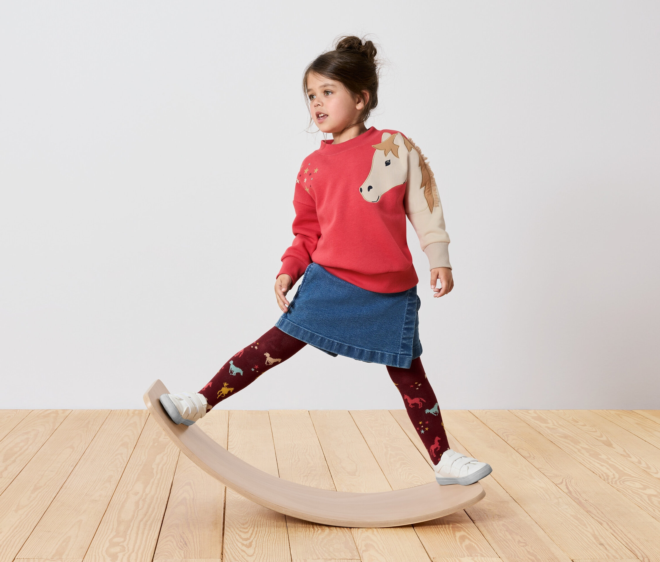 Une fille en pull rouge à motif de cheval, jupe en jean et collant à motifs se tient sur une planche d'équilibre en bois.