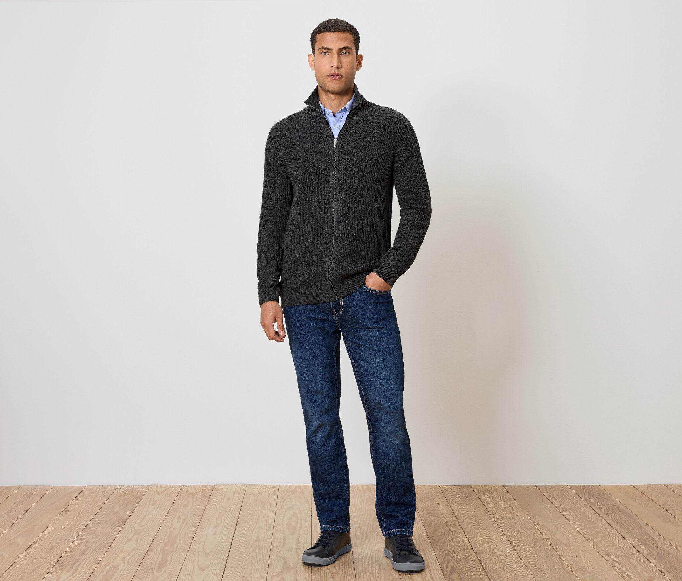 Un homme debout porte une veste en maille anthracite chiné, un jean et des chaussures.