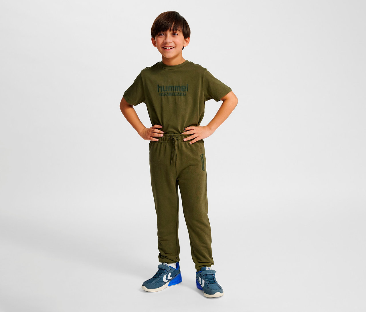 Garçon debout, les mains sur les hanches, portant un ensemble vert avec un pantalon régulier Hummel HMLJR Base.