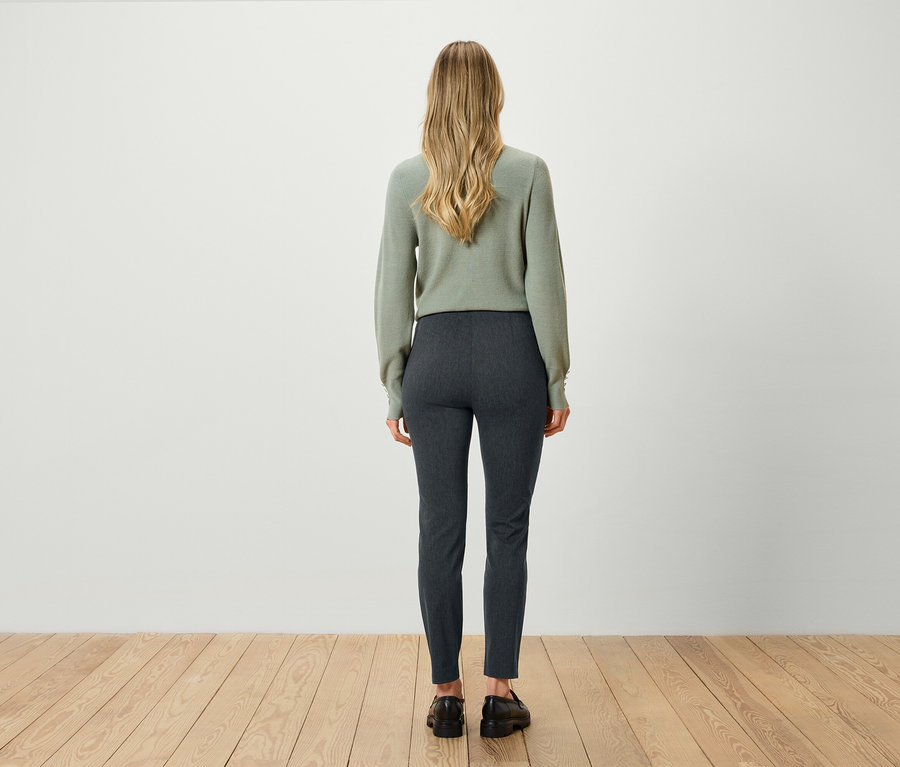 Vue arrière d'une femme blonde portant un pull vert et un pantalon gris.