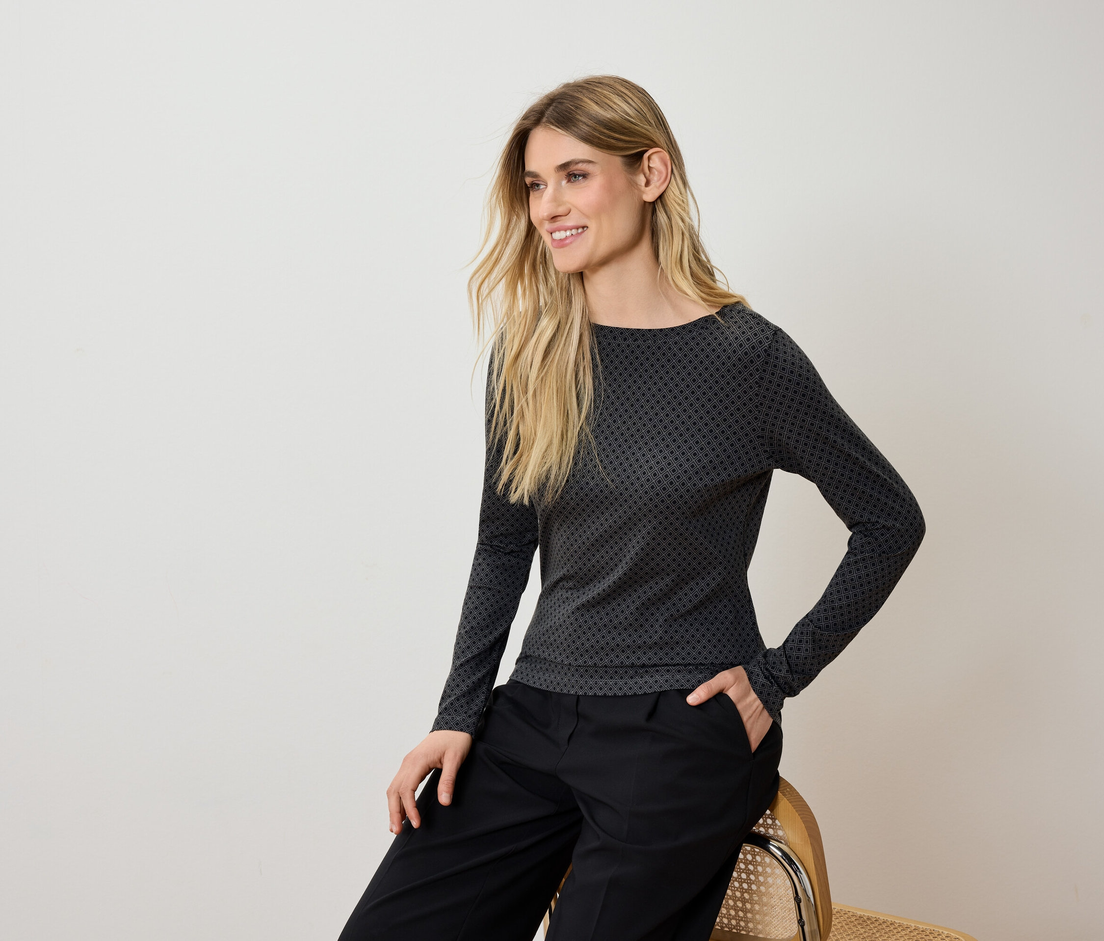 Une femme blonde est assise sur une chaise, portant un pantalon noir et un t-shirt à manches longues gris et noir.