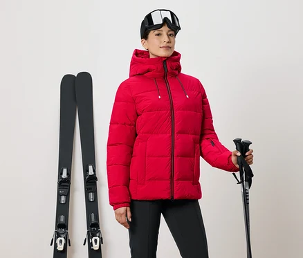 Femme en veste de ski rouge tenant des bâtons, skis à côté et lunettes de ski sur la tête.