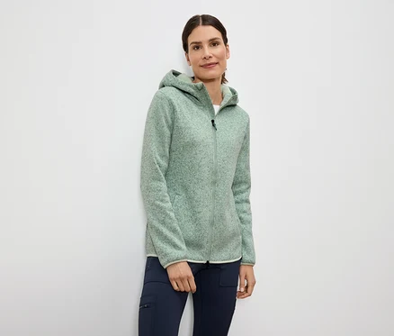 Une femme porte une veste polaire vert clair zippée à capuche et un pantalon bleu marine.