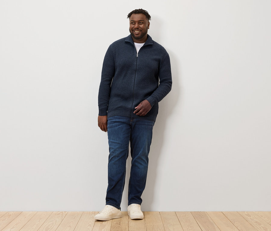 Un homme debout portant une veste en maille bleu marine chiné, un jean et des baskets.