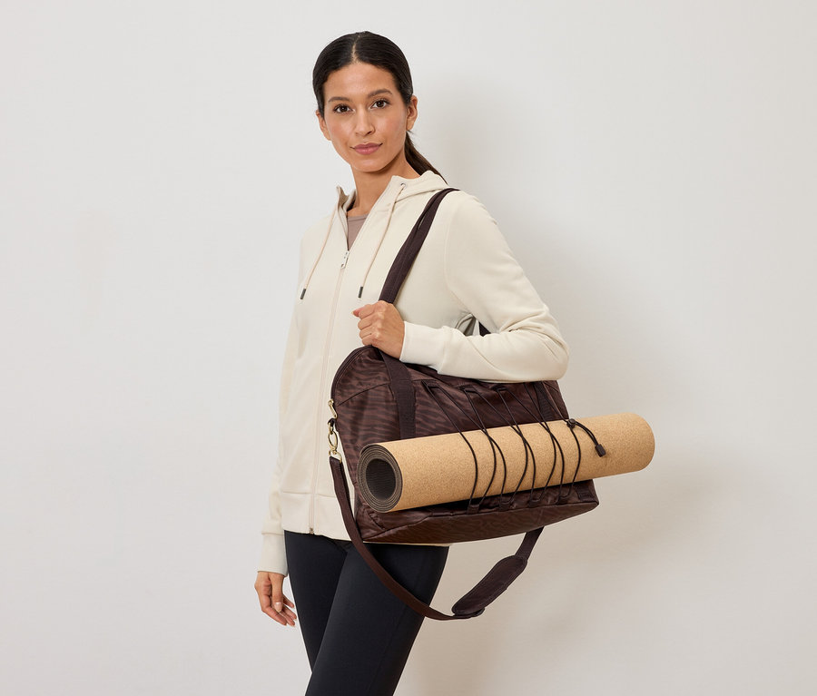 Femme portant un sac de sport marron avec un tapis de yoga sur l'épaule, une veste crème et un legging noir.