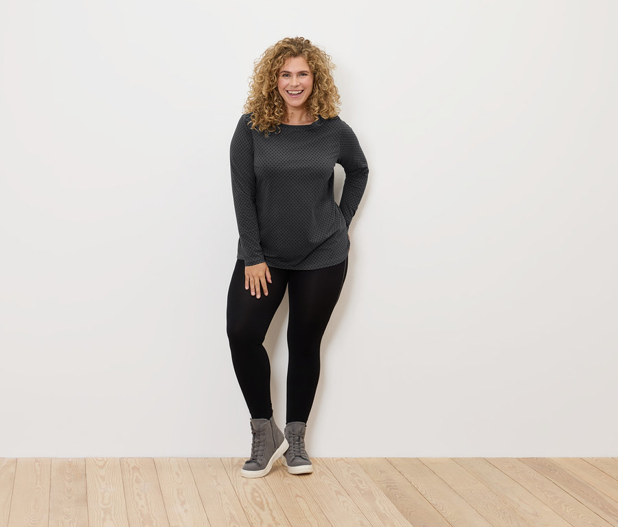 Une femme aux cheveux bouclés se tient debout et sourit, elle porte des leggings noirs, des chaussures grises et un t-shirt gris et noir.