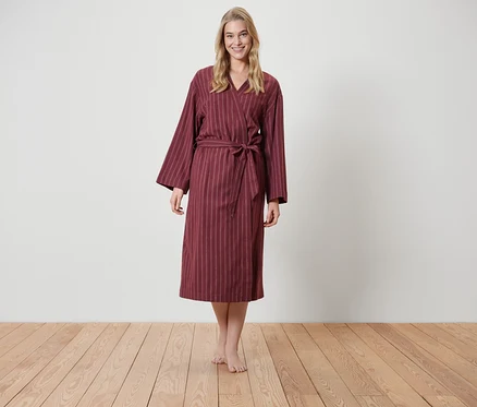 Une femme blonde se tient sur un plancher en bois et porte un peignoir bordeaux rayé.
