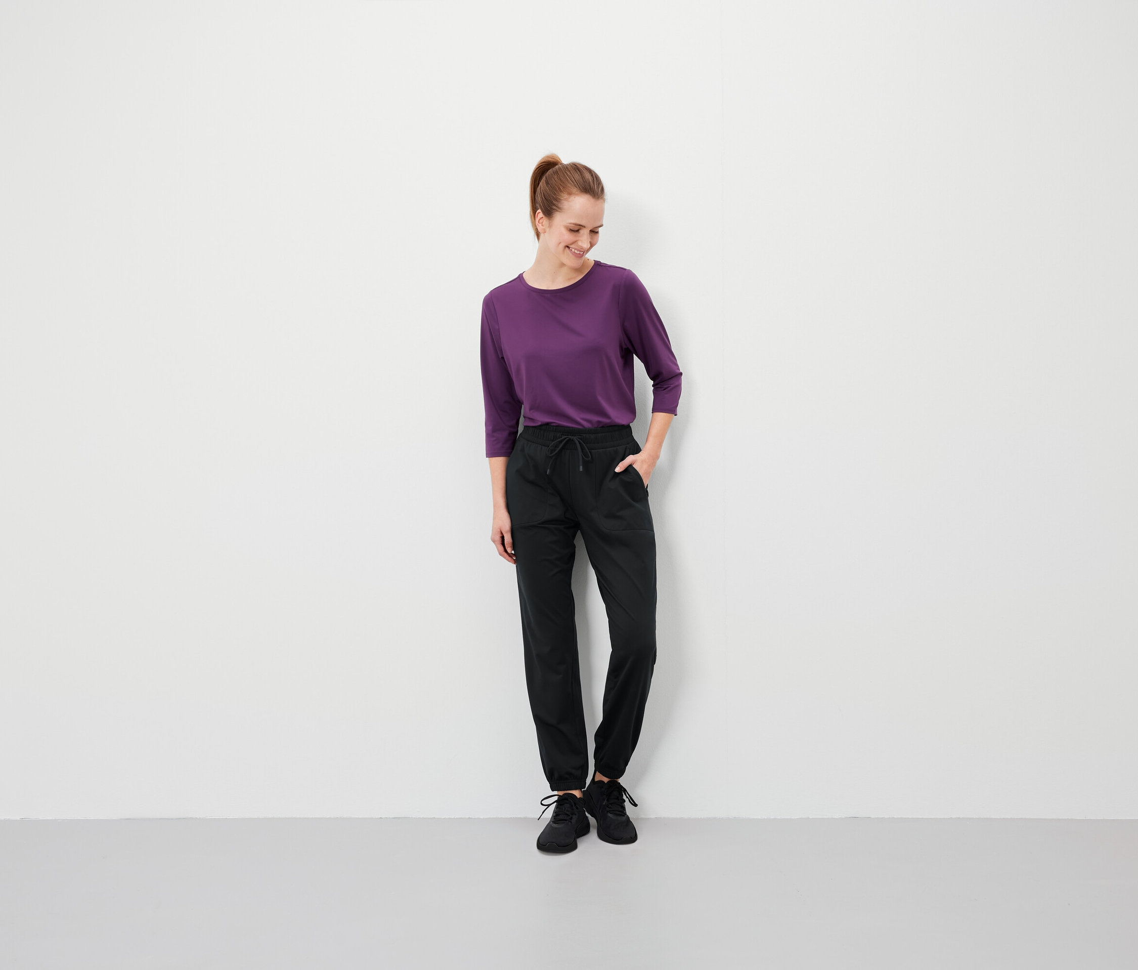 Une femme aux cheveux bruns, coiffés en queue de cheval, se tient debout, les mains dans les poches de son pantalon noir, et porte un T-shirt violet.