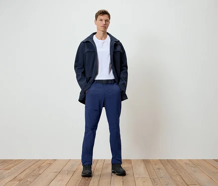 Un homme se tient sur un plancher en bois. Il porte une veste bleu foncé, un t-shirt blanc, un pantalon bleu et des chaussures noires.