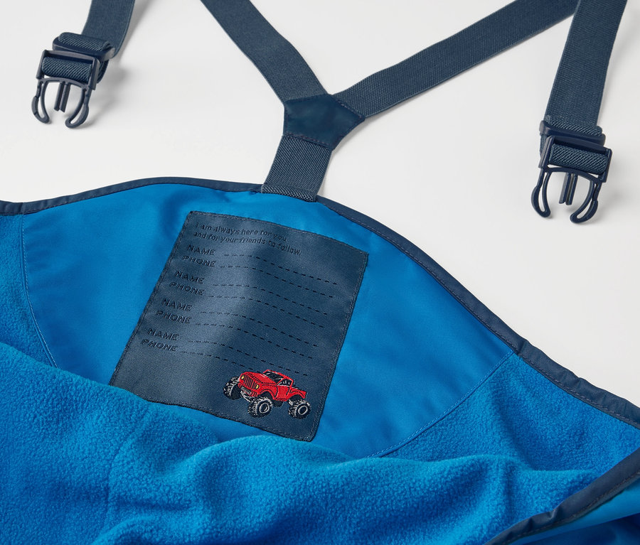 Gros plan sur un pantalon de pluie thermique bleu pour enfant avec bretelles et un patch de voiture rouge.