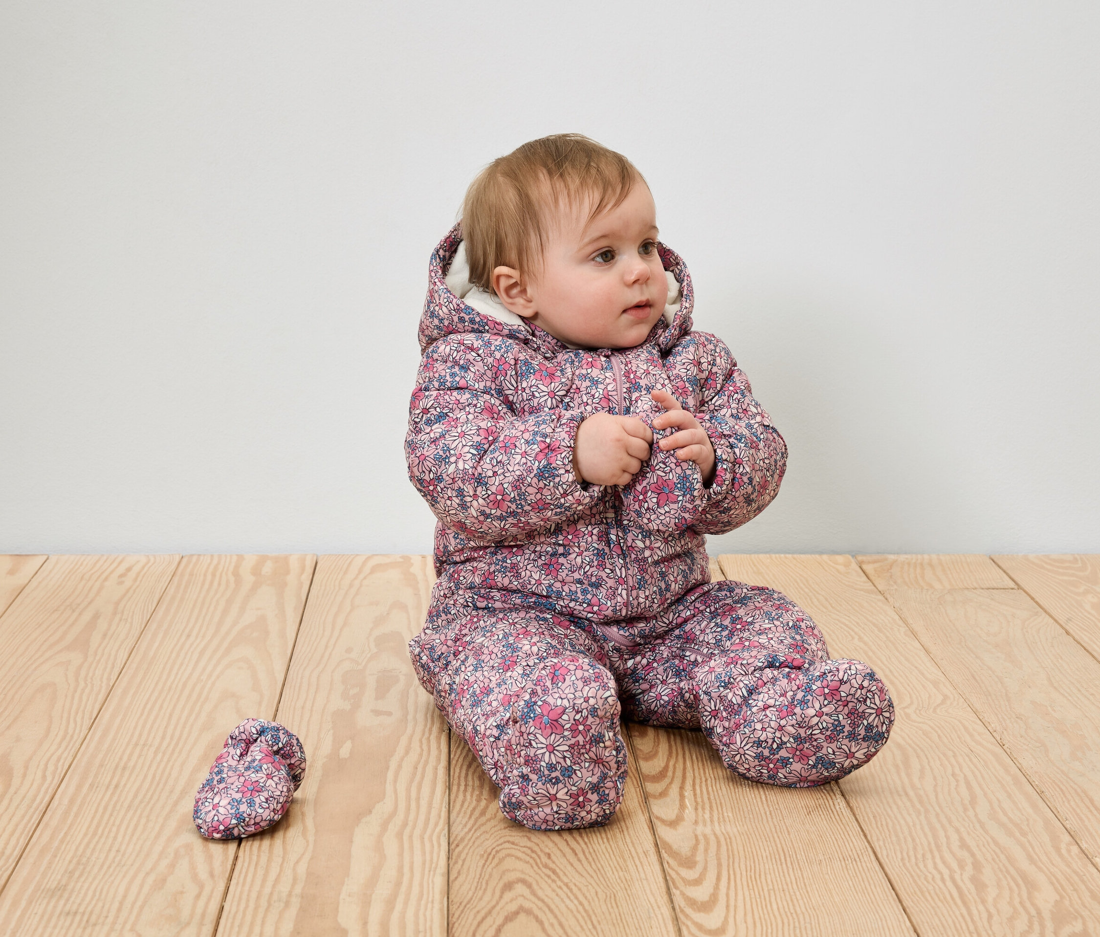 Une petite fille est assise sur un plancher de bois dans une combinaison d'hiver fleurie à capuche avec une botte.