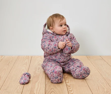 Une petite fille est assise sur un plancher de bois dans une combinaison d'hiver fleurie à capuche avec une botte.