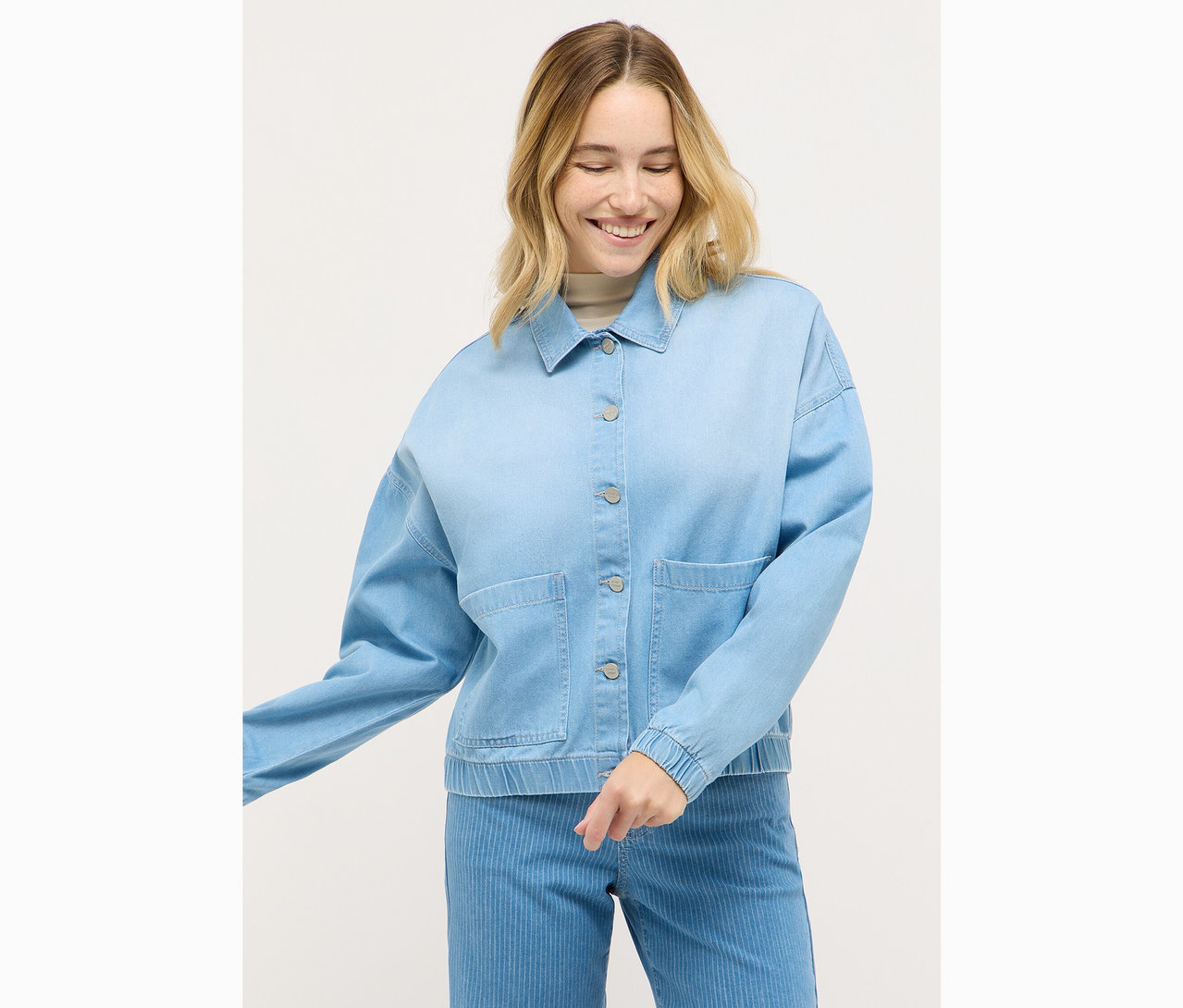 Une femme blonde souriante porte une veste en jean bleu clair boutonnée avec des boutons argentés et un pantalon bleu clair.