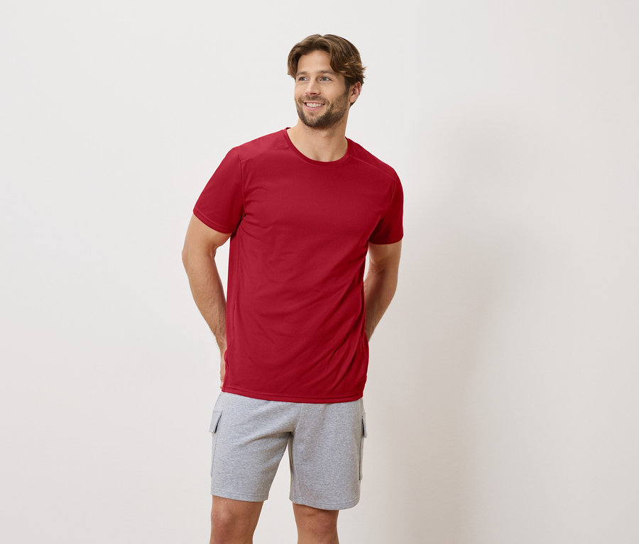 Un homme se tient debout, les mains derrière le dos, portant un t-shirt rouge et un short gris.