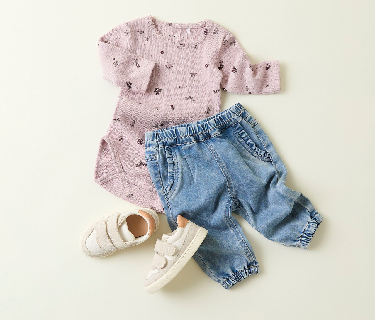 Un body rose à fleurs, un jean pour bébé NAME IT et des chaussures beige sont posés sur un fond clair.