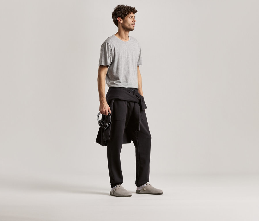 Un homme porte un t-shirt gris et un pantalon de survêtement noir. Il tient un casque audio dans sa main.