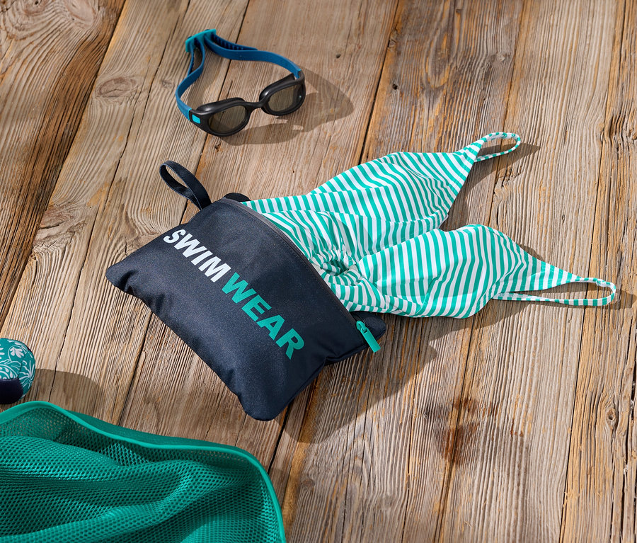 Photo d'accessoires de natation sur une surface en bois, comprenant un sac avec l'inscription « Swimwear », un maillot de bain rayé vert et blanc, des lunettes de natation et un filet vert.